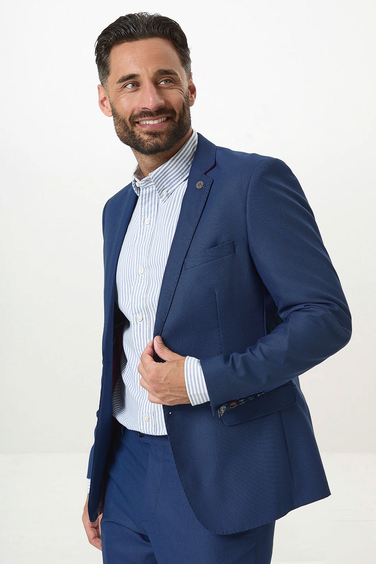 A man with dark hair and a beard smiles while wearing the Marc Darcy DANNY - Royal Blue Two Piece Suit, paired with a light blue striped shirt. He holds his slim fit suit jacket lapel, standing against a white background.