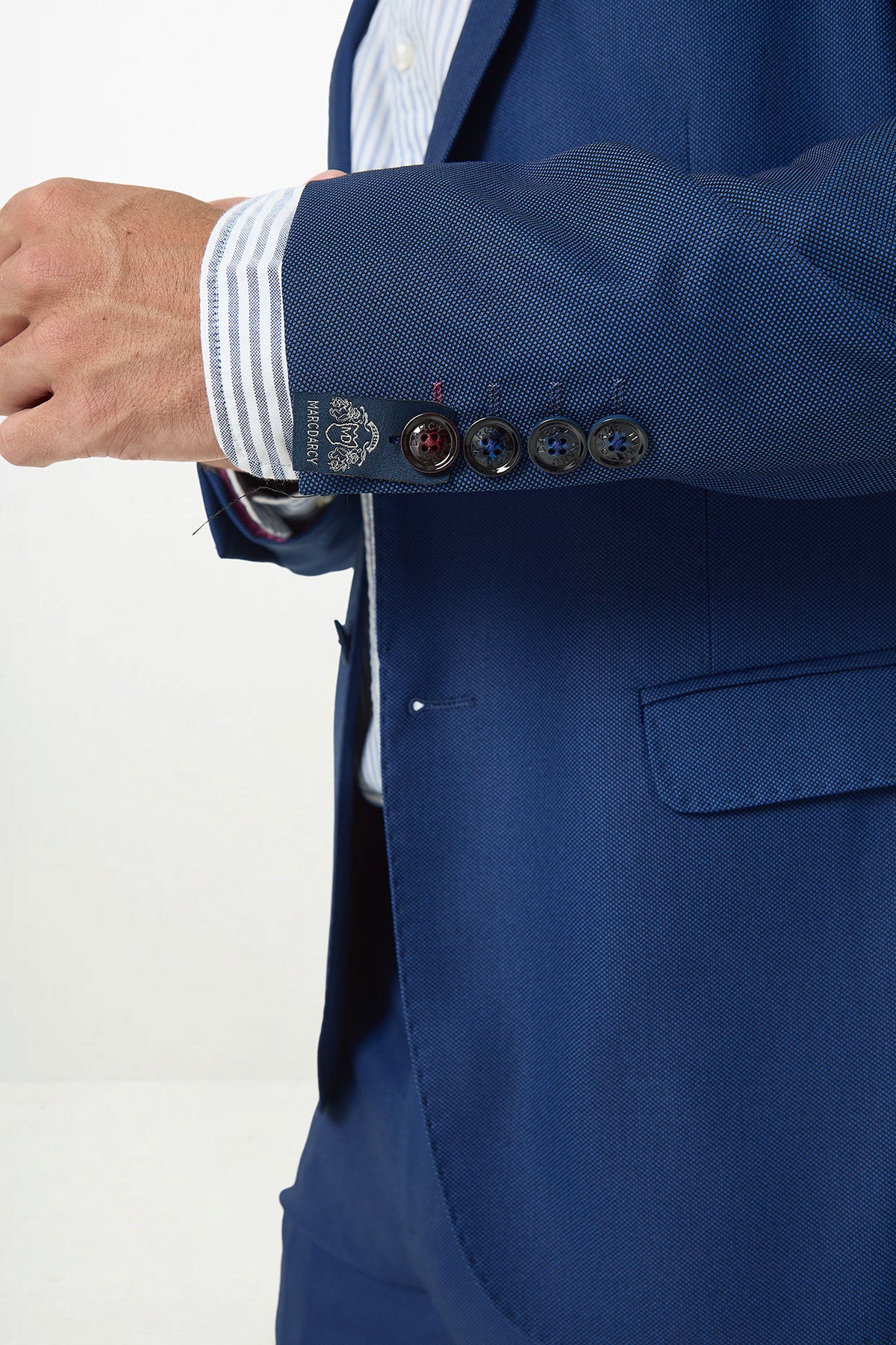 A person wearing the Marc Darcy DANNY Royal Blue Two Piece Suit, featuring visible sleeve buttons, and a blue and white striped shirt, holds their left arm bent at the elbow against a plain background.