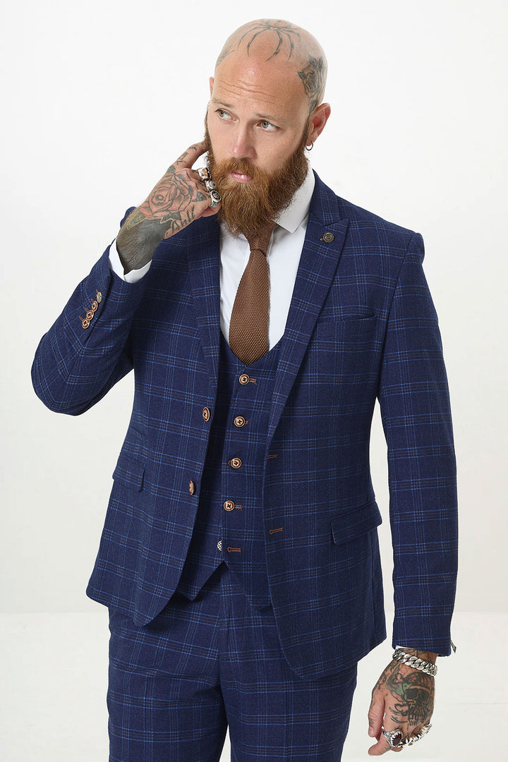 A bearded, tattooed man in the marcdarcy CHIGWELL Blue Tweed Check Three Piece Suit with a brown tie and white shirt stands against a plain background, touching his neck and looking to the side.