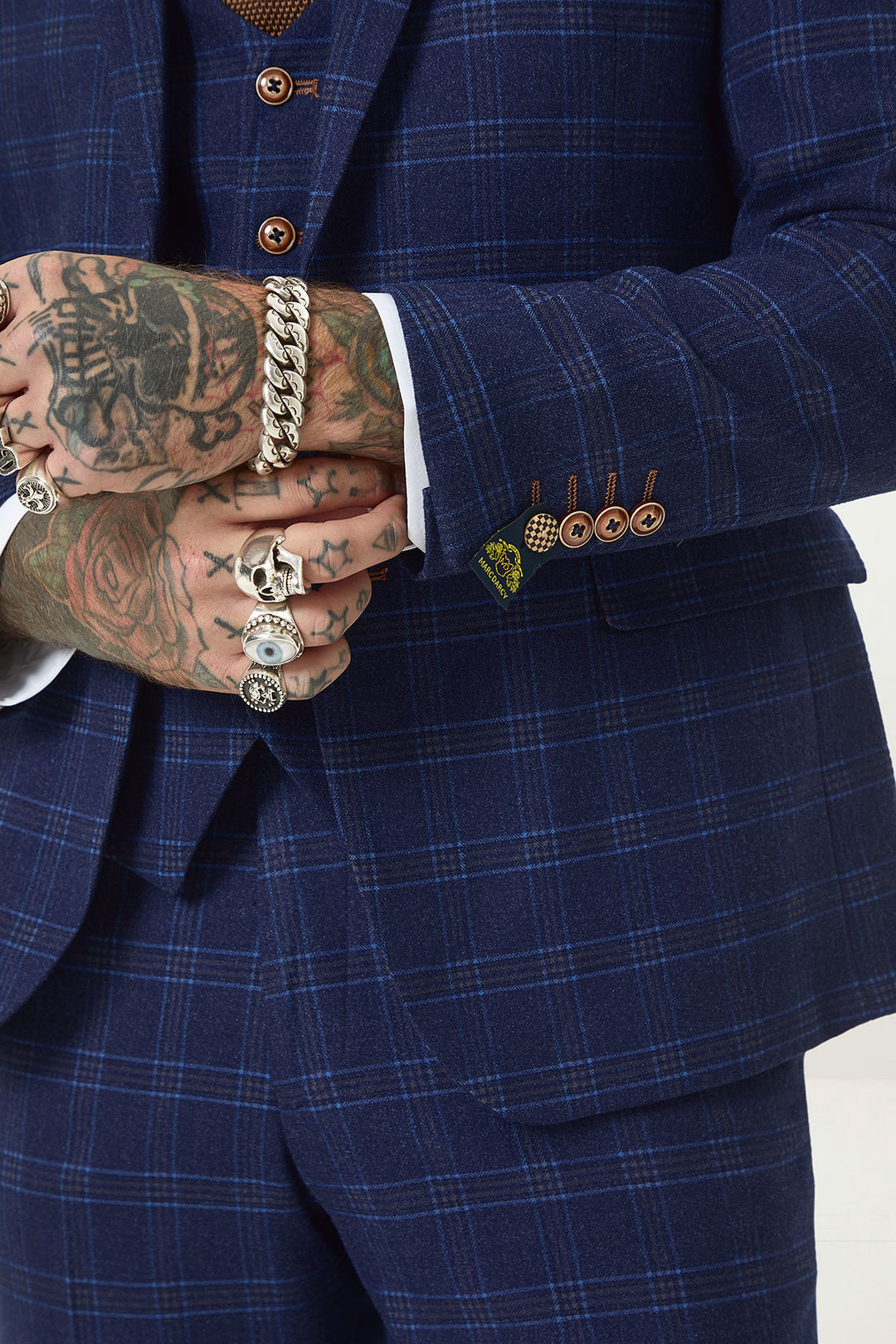 A tattooed individual wearing multiple rings and a chain bracelet adjusts the sleeve of their marcdarcy CHIGWELL Blue Tweed Check Three Piece Suit, showcasing its slim fit, brown buttons, and visible vest underneath.