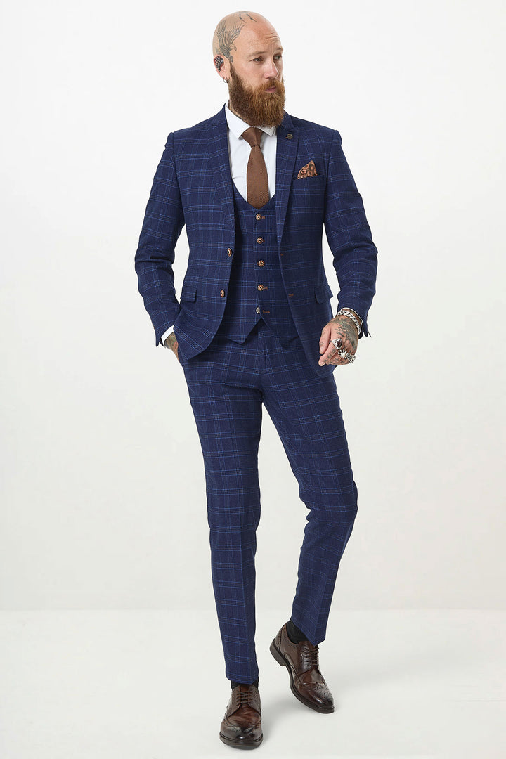 A bearded, tattooed man models the marcdarcy CHIGWELL Blue Tweed Check Three Piece Suit with a white shirt, brown tie, and brown shoes. He stands confidently with one hand in his pocket against a plain white background.