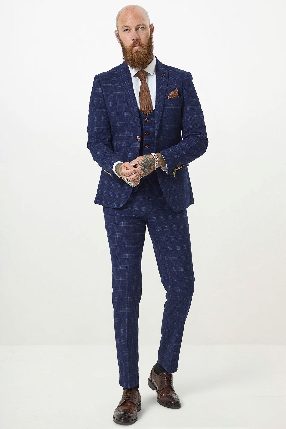 A bearded man models the marcdarcy CHIGWELL Blue Tweed Check Three Piece Suit in slim fit with a white shirt, brown tie, and brown dress shoes. His tattooed hands stand out against a plain white background—perfect for a stylish wedding guest look.
