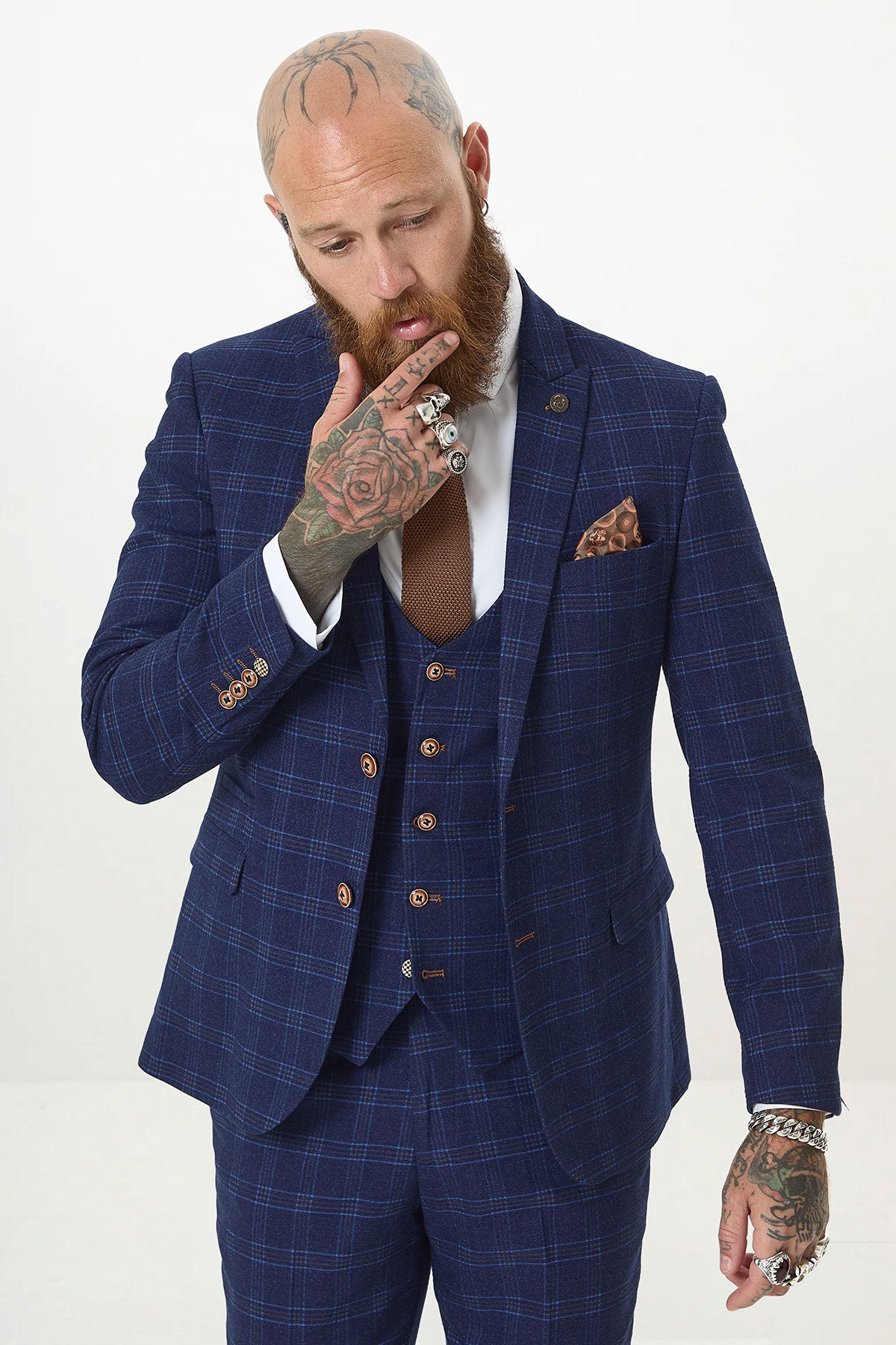 A tattooed man in the marcdarcy CHIGWELL Blue Tweed Check Three Piece Suit, paired with a white shirt and brown tie, poses thoughtfully with his hand to his chin.