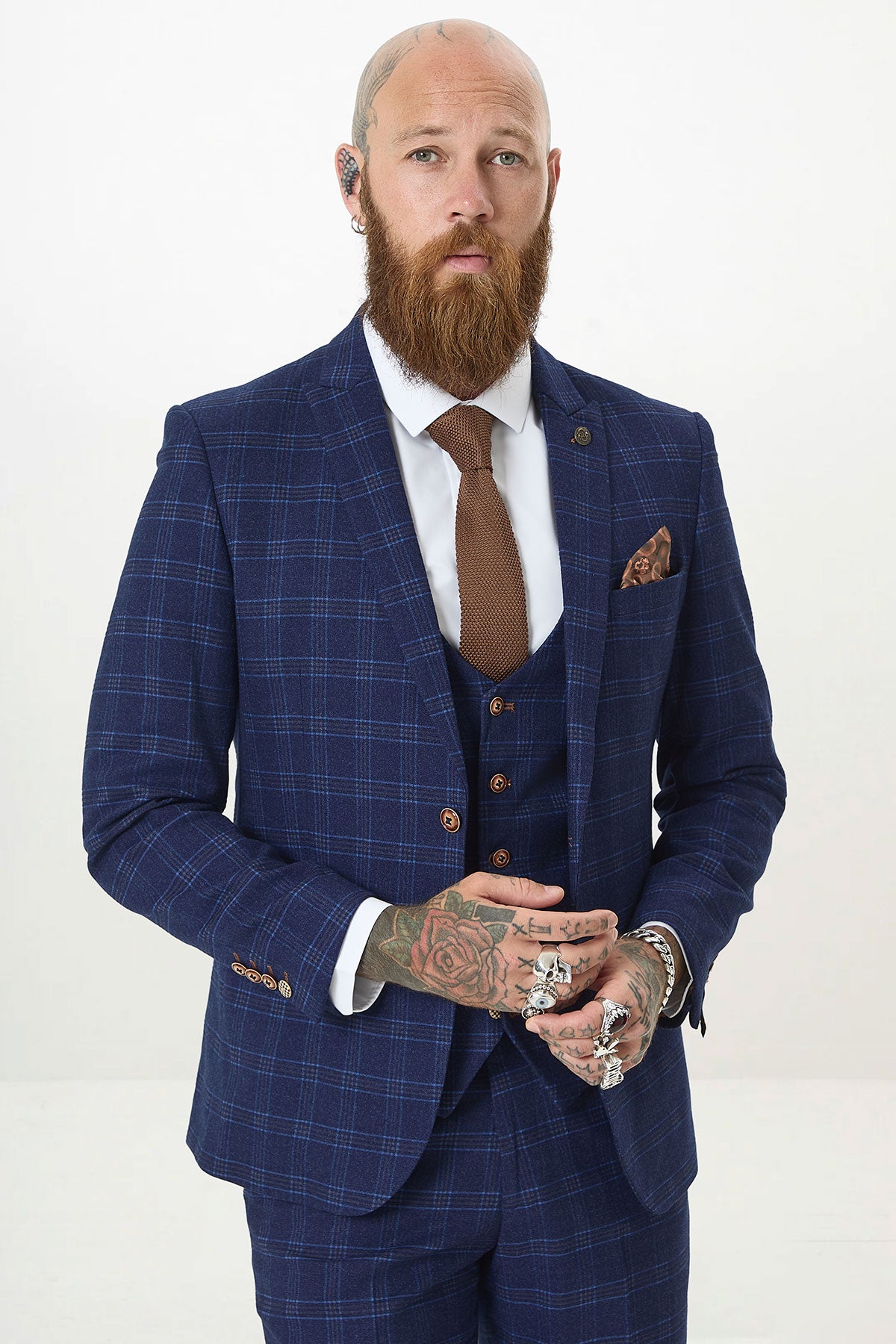 A bearded man with hand tattoos confidently wears the marcdarcy CHIGWELL Blue Tweed Check Blazer, a white shirt, brown tie, multiple rings, pocket square, and an earpiece against a plain white background.