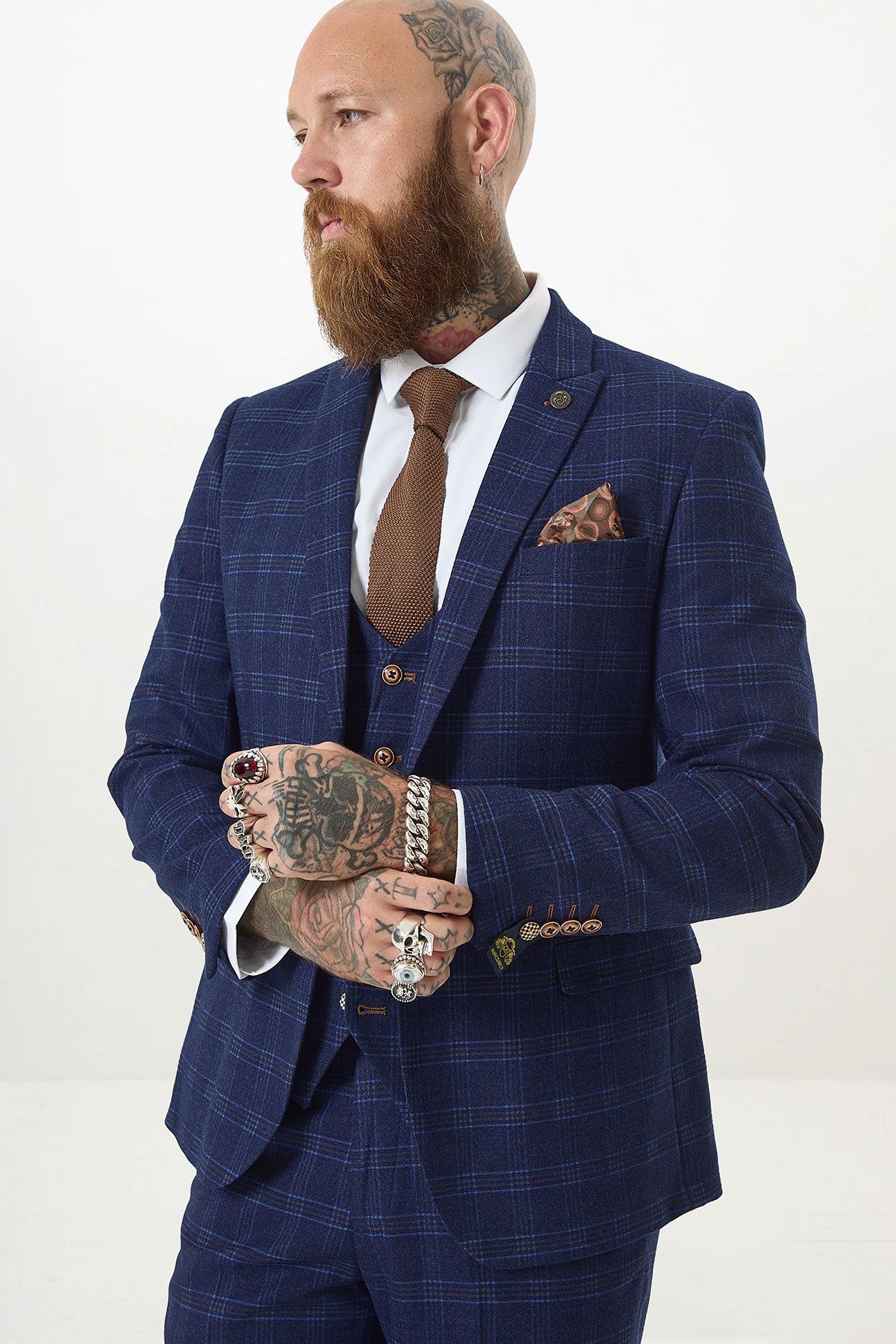 A bearded, tattooed man with rings wears the marcdarcy CHIGWELL Blue Tweed Check Blazer, styled with a brown tie and pocket square. He stands against a plain white background, looking sideways with his hands clasped.