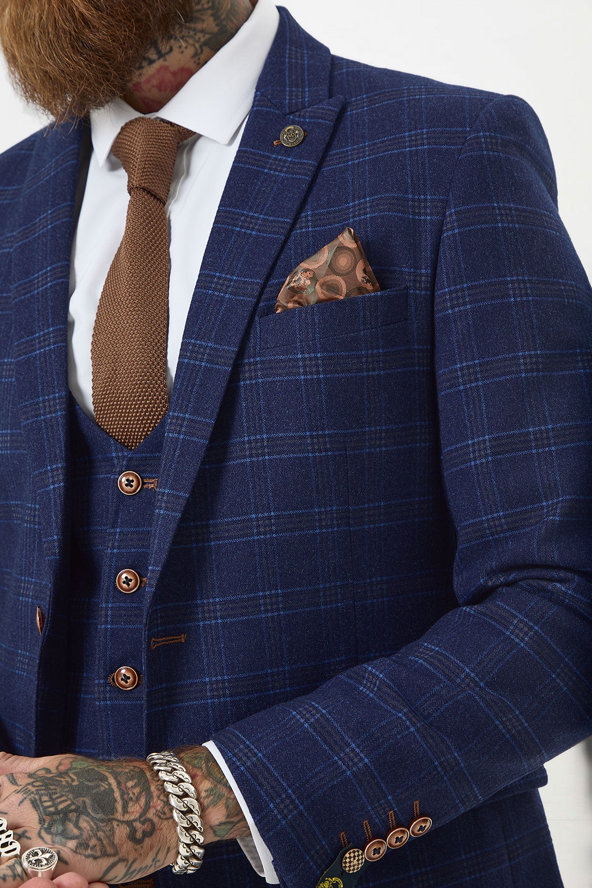 A person wears the marcdarcy CHIGWELL Blue Tweed Check Blazer in a slim fit, styled with a brown tie, white shirt, patterned pocket square, and visible silver jewelry over tattooed hands; the blazer features brown buttons and stitching accents.
