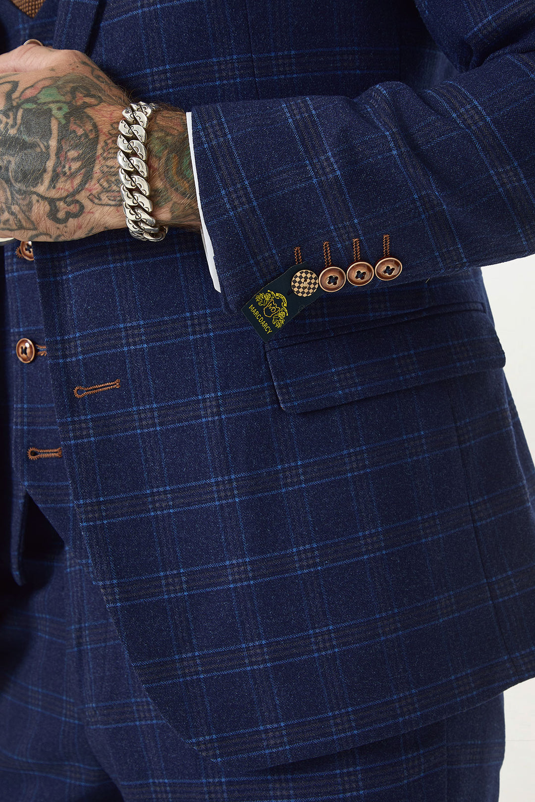 A person wearing the marcdarcy CHIGWELL Blue Tweed Check Blazer with brown buttons and a visible sleeve label, displaying tattooed hands and a thick silver bracelet.