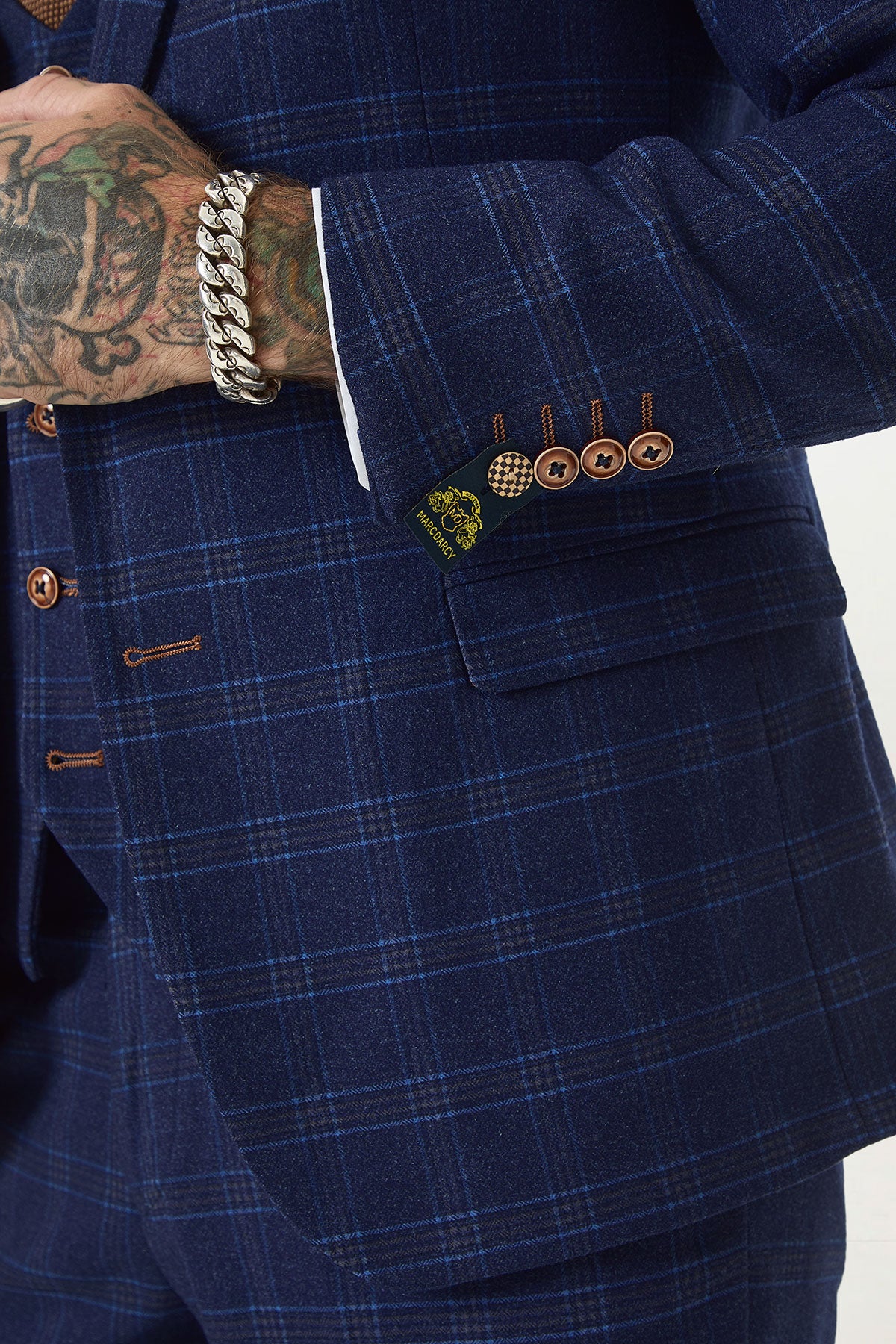 A person wears the marcdarcy CHIGWELL - Blue Tweed Check Blazer with brown buttons, revealing a tattooed hand and chunky silver bracelet. The sleeve label displays the brand's yellow and black logo, highlighting this stylish men's blazer.