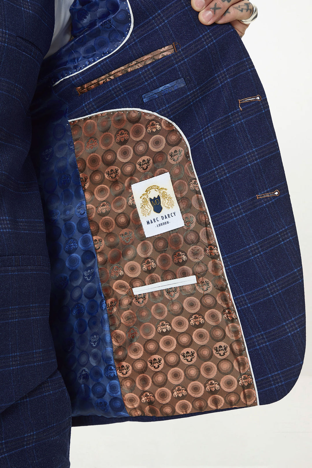 A close-up of a person holding open the marcdarcy CHIGWELL Blue Tweed Check Blazer, revealing its inner lining with a brown circular pattern and a marcdarcy label sewn onto the pocket.