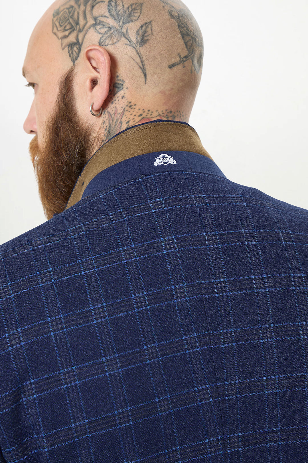 A bearded person with a floral head tattoo and earring is shown from behind wearing the marcdarcy CHIGWELL Blue Tweed Check Blazer, featuring a brown collar and a small white embroidered logo on the back.