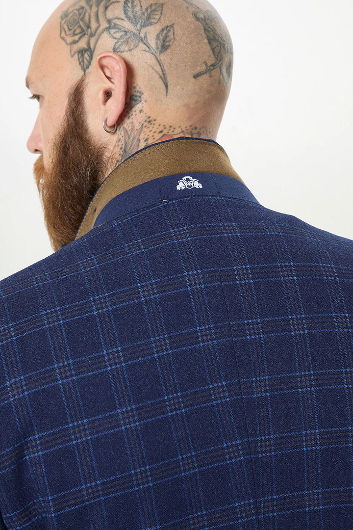A bearded person with a floral head tattoo and earring is shown from behind wearing the marcdarcy CHIGWELL Blue Tweed Check Blazer, featuring a brown collar and a small white embroidered logo on the back.
