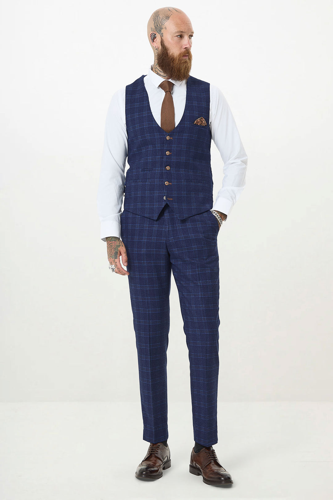 A bearded, tattooed man wears the marcdarcy CHIGWELL Blue Tweed Check Three Piece Suit with a white shirt, brown tie, pocket square, and brown dress shoes against a plain white background.