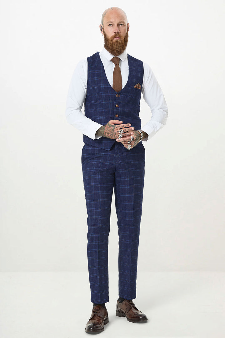 A bearded, tattooed man wears the marcdarcy CHIGWELL Blue Tweed Check Waistcoat with a white shirt and brown tie, standing against a plain light background and looking at the camera with his hands together.