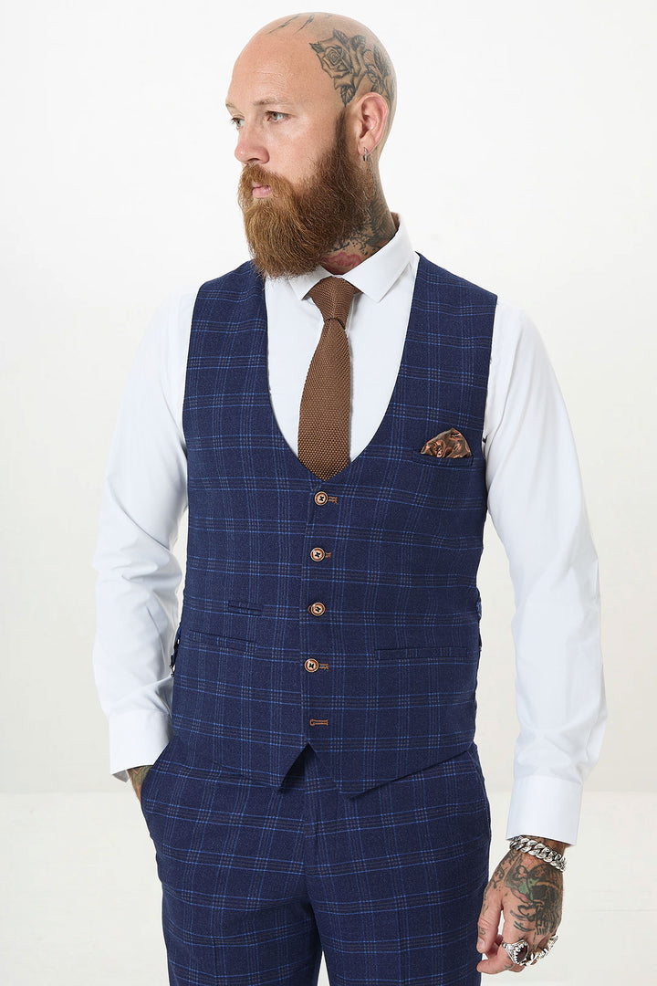A bearded man with tattoos wears the marcdarcy CHIGWELL Blue Tweed Check Waistcoat, paired with a white dress shirt, brown tie, and pocket square. He stands hands in pockets, looking to the side against a plain background.