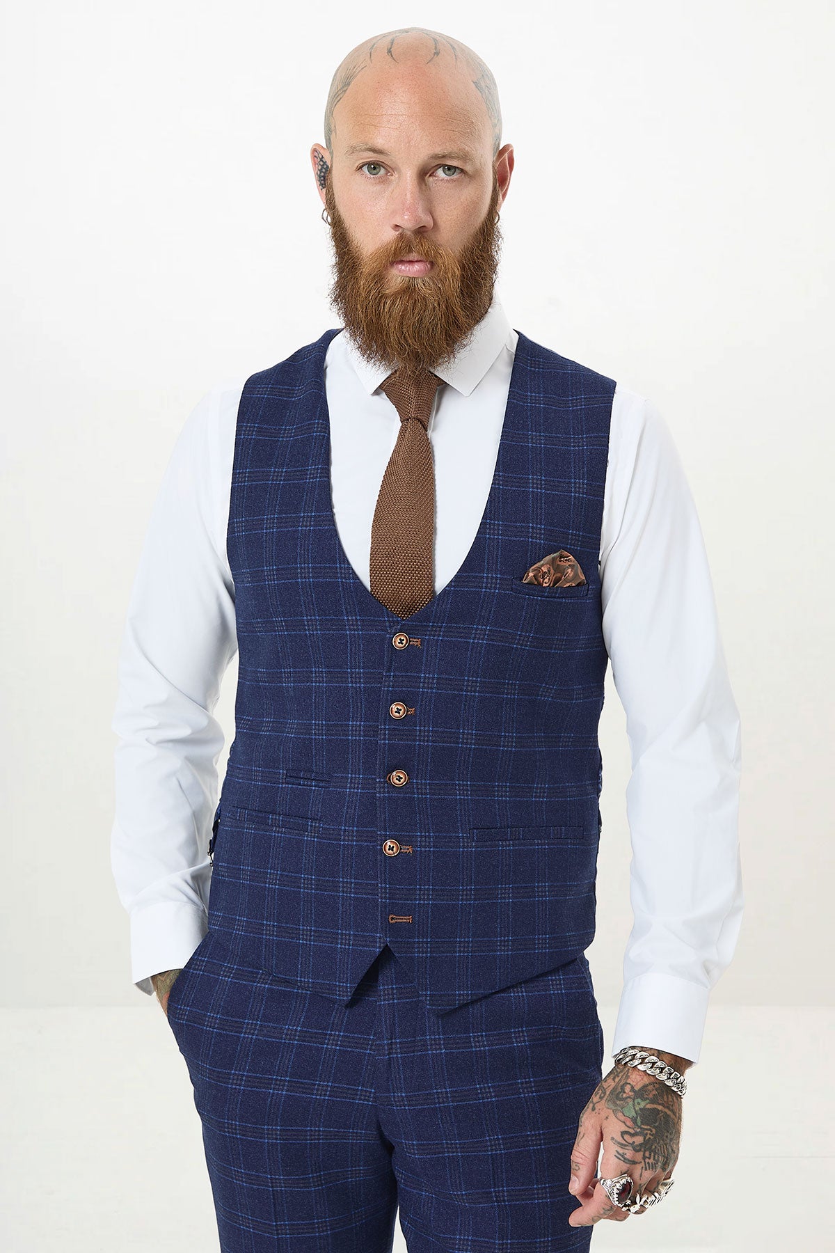 A bearded man with tattoos, wearing a white shirt, brown tie, and marcdarcy CHIGWELL - Blue Tweed Check Waistcoat with matching slim fit pants, stands with one hand in his pocket against a plain light background.