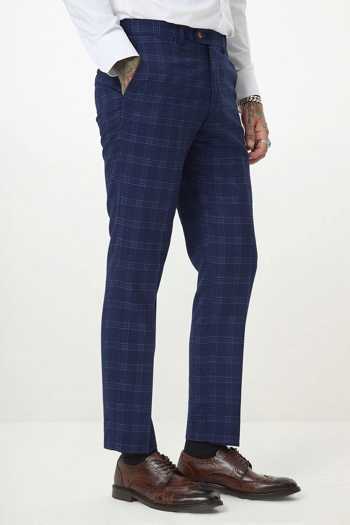 Wearing marcdarcy’s CHIGWELL Blue Tweed Check Trousers with a white shirt and brown leather dress shoes, a person stands with one hand in their pocket, showing tattooed hands. The photo is cropped at the torso and ankles.