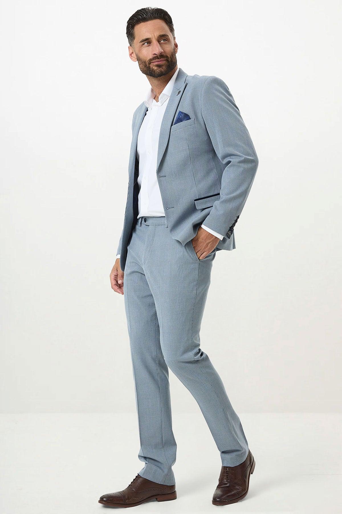 A man wears the Marc Darcy BROMLEY Sky Blue Check Two Piece Suit with a white shirt and brown dress shoes, hand in pocket, looking to the side. An ideal choice for a stylish men’s wedding suit.