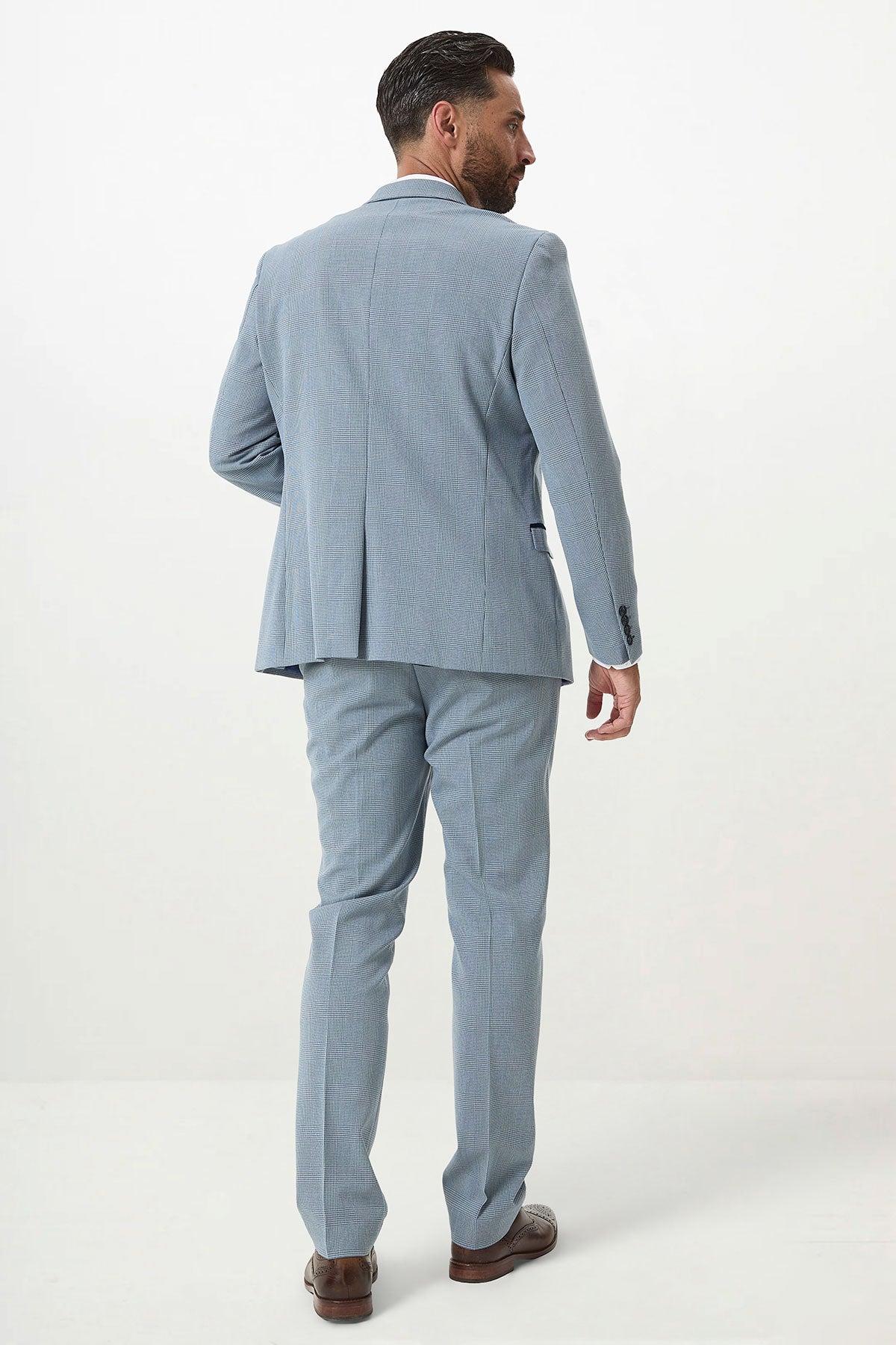 A man in the Marc Darcy BROMLEY Sky Blue Check Two Piece Suit with brown shoes stands facing away from the camera in a minimalistic white setting.
