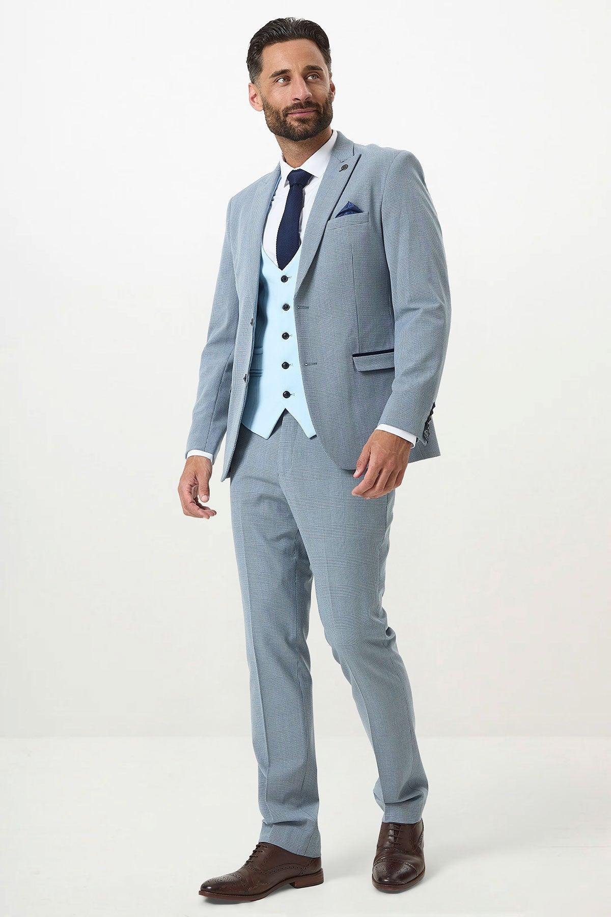 A man with short dark hair and a beard stands against a plain background wearing the Marc Darcy BROMLEY - Sky Check Suit with Kelvin Sky Waistcoat, white shirt, navy tie, pocket square, and brown dress shoes.