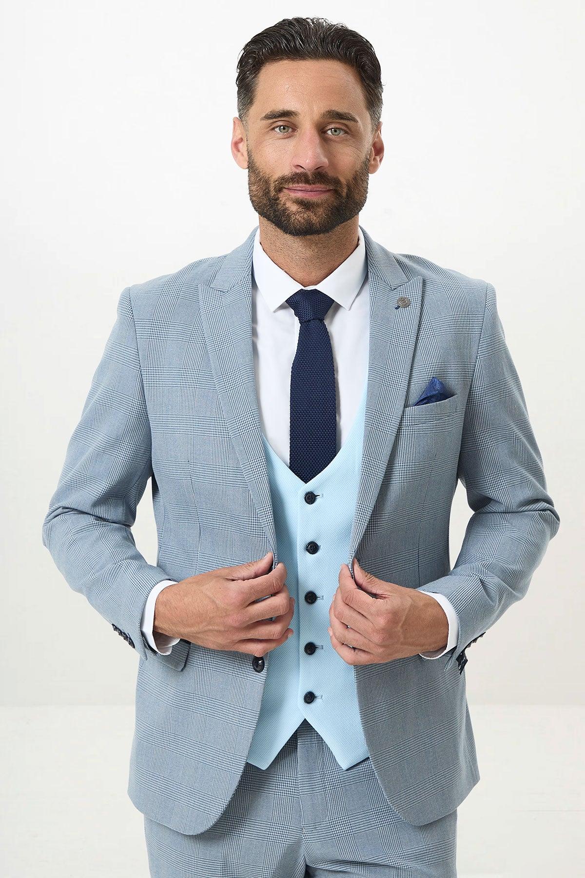 A man with dark hair and a beard wears the Marc Darcy BROMLEY Sky Check Suit with Kelvin Sky Waistcoat, paired with a white shirt and dark blue tie. He adjusts his jacket while standing against a plain white background, looking at the camera.