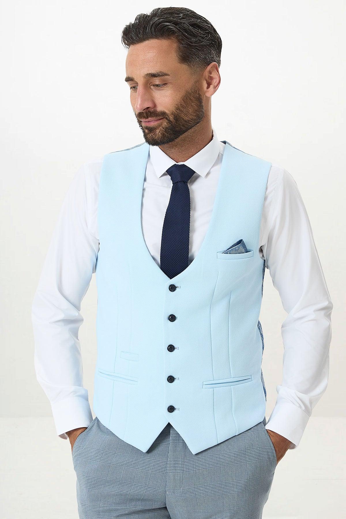 A man with dark hair and a beard wears the marcdarcy KELVIN Sky Blue Single Breasted Waistcoat over a white shirt, paired with a navy tie and grey checkered pants. He stands with hands in pockets, looking slightly down and to the side.