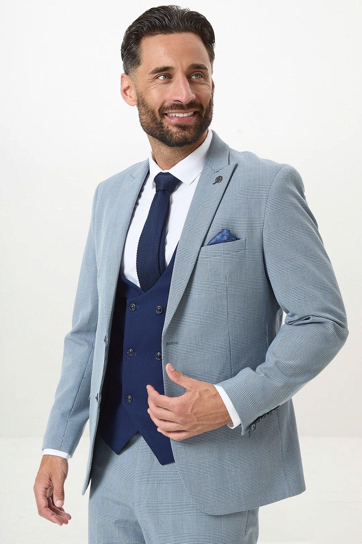 A man with dark hair and a beard wears the Marc Darcy BROMLEY Sky Check Suit with Kelvin Royal Waistcoat, paired with a white shirt and dark blue tie. He is smiling and posing against a plain light background.
