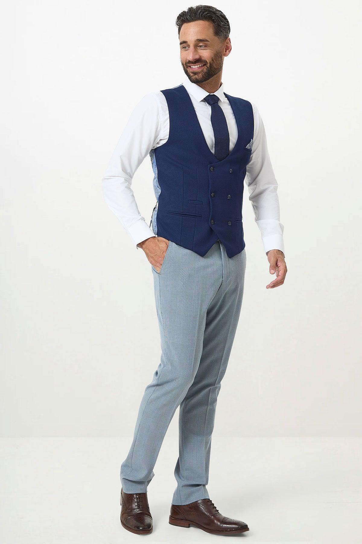A man wearing a white dress shirt, marcdarcy KELVIN Royal Blue Double Breasted Waistcoat, blue tie, light gray trousers, and brown dress shoes stands smiling and looking to his left against a plain white background.