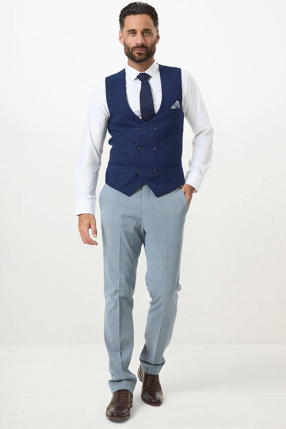 A man wearing a white dress shirt, marcdarcy KELVIN royal blue double breasted waistcoat, navy tie, light gray dress pants, and brown dress shoes stands against a plain white background.
