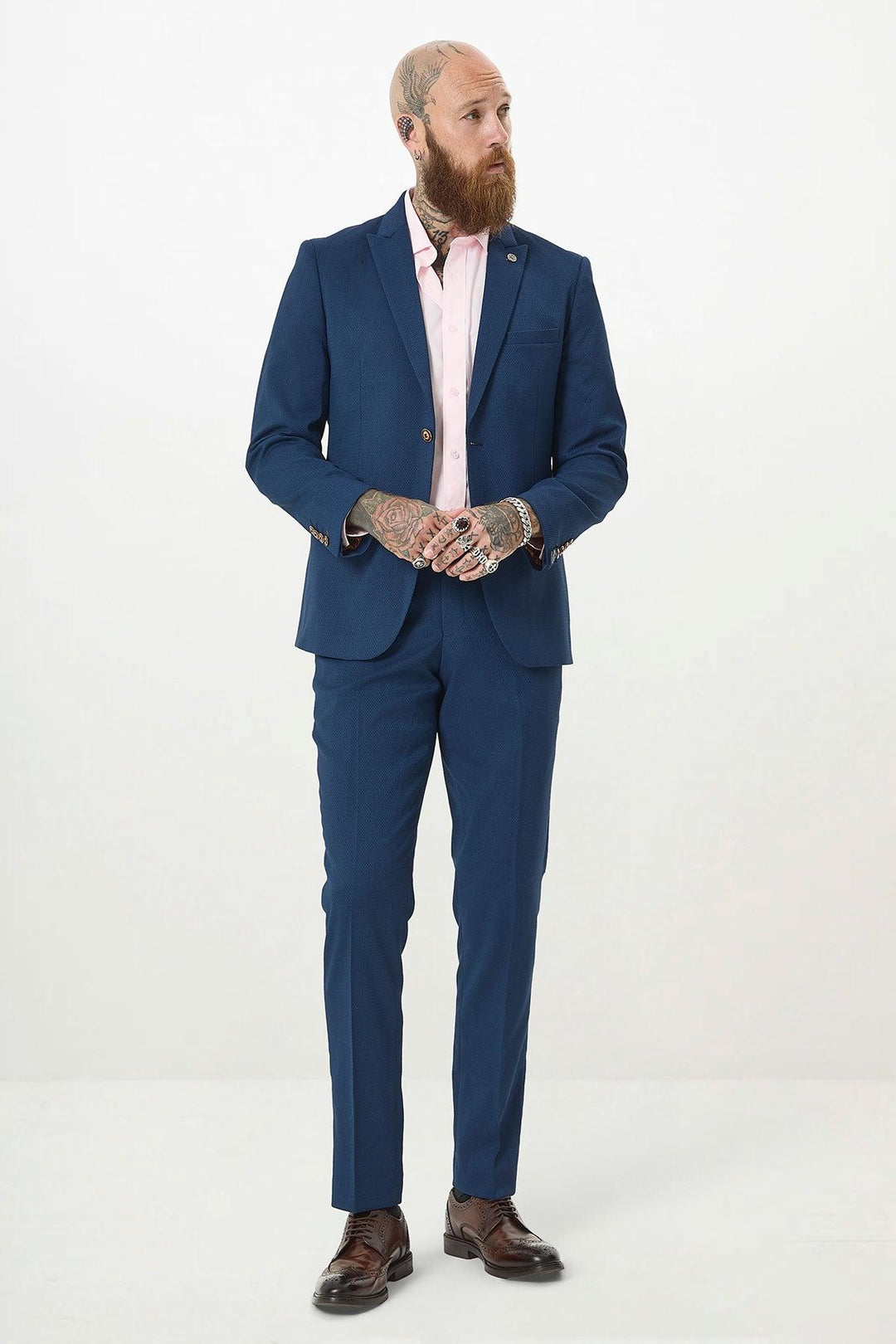 A bearded, tattooed man in a MAX - Royal Blue Two Piece Suit by Marc Darcy with a light pink shirt and brown dress shoes stands against a white background, looking left with hands clasped.