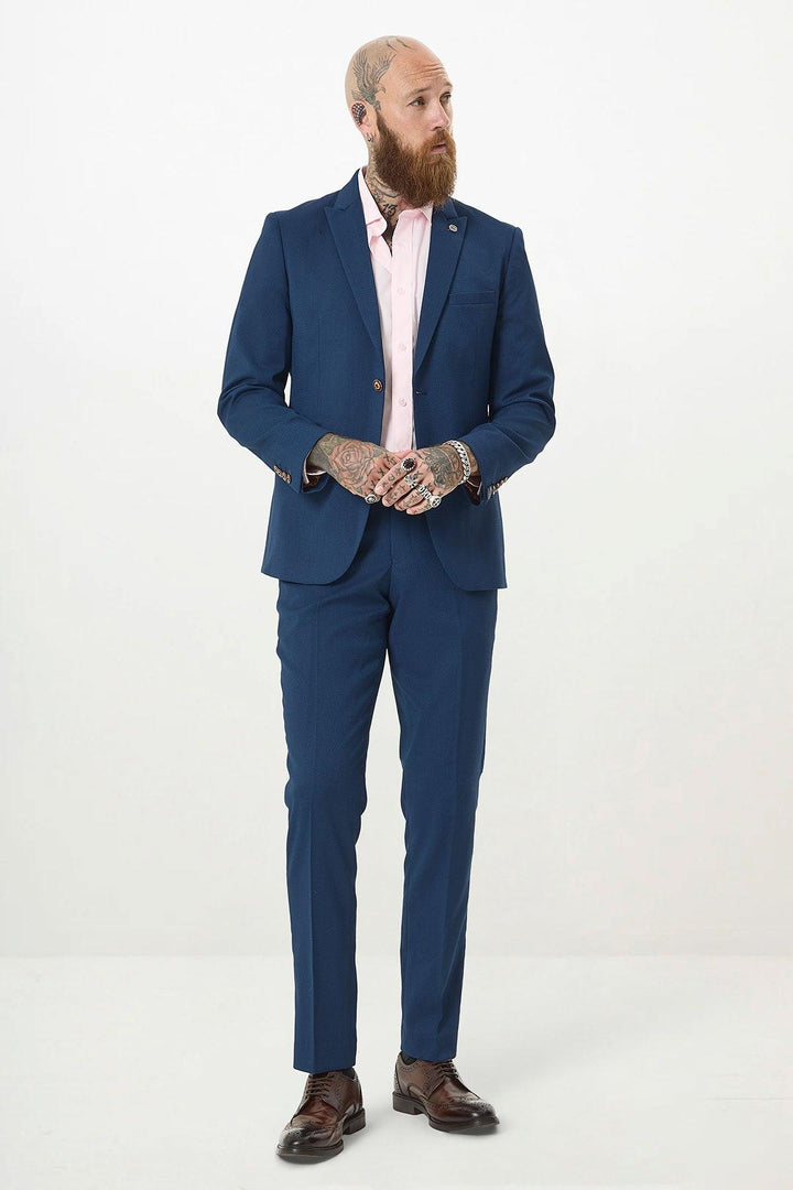 A bearded, tattooed man in a MAX - Royal Blue Two Piece Suit by Marc Darcy with a light pink shirt and brown dress shoes stands against a white background, looking left with hands clasped.