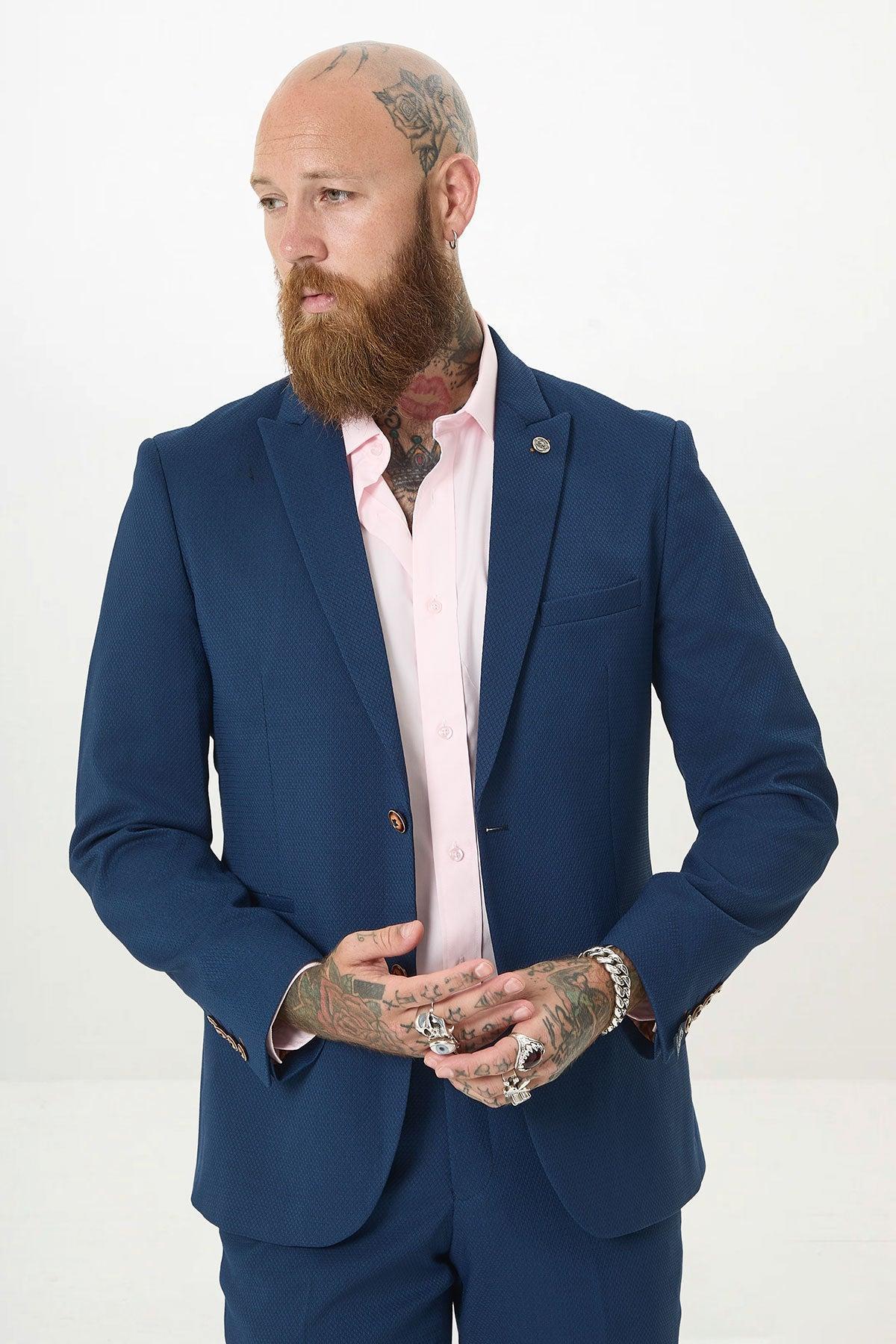 A bearded, tattooed, bald man wears the marcdarcy MAX - Royal Blue Blazer with Contrast Buttons over a light pink shirt. He looks left with hands clasped against a white background, ideal for office wear.