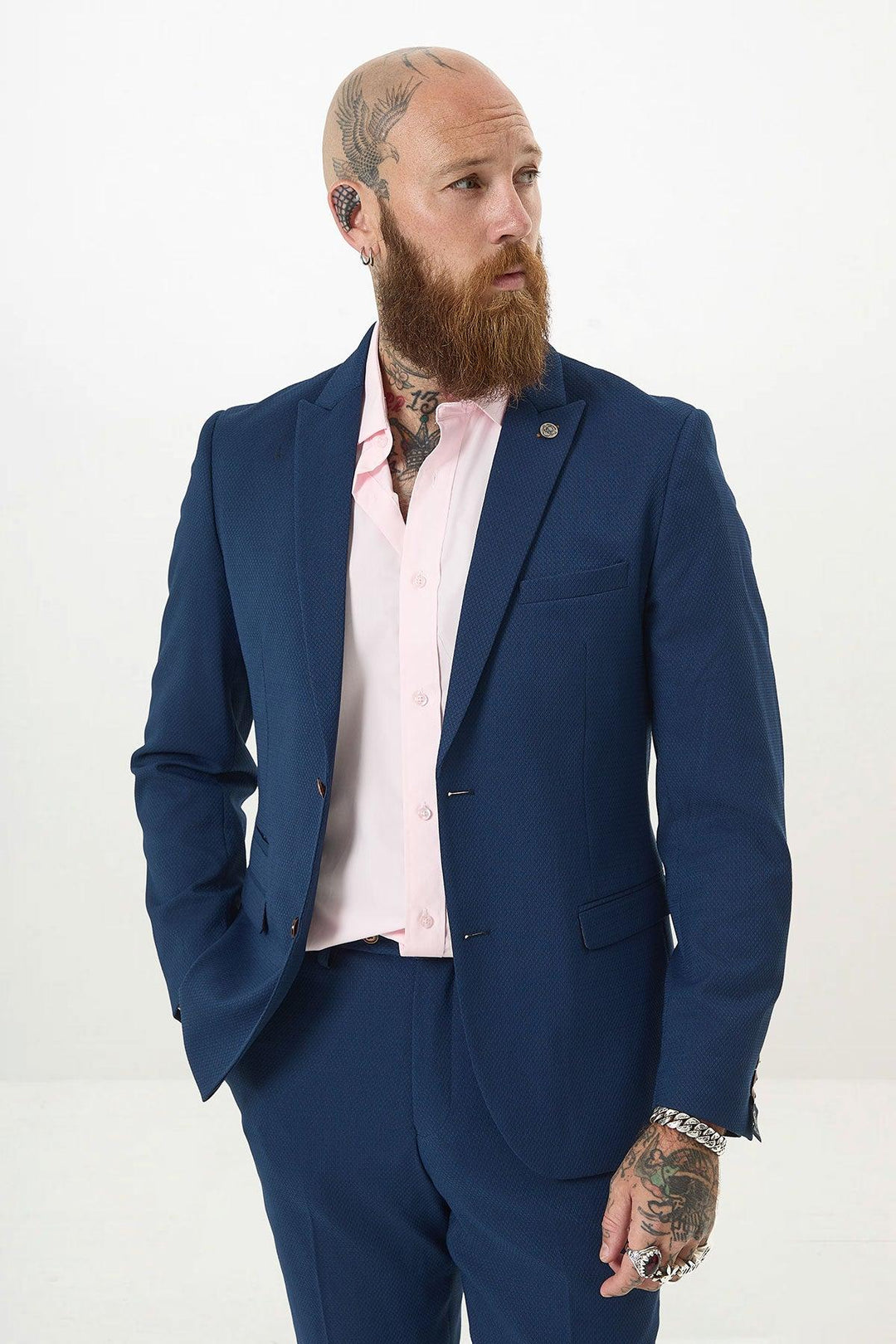 A bearded man with tattoos, dressed in the Marc Darcy MAX - Royal Blue Two Piece Suit and a light pink shirt, stands with one hand in his pocket, looking to the side against a plain white background.