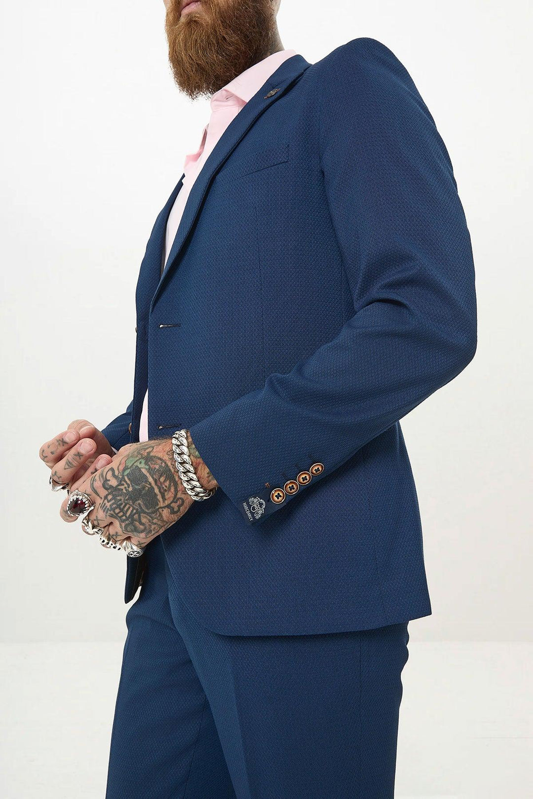A man with a beard and tattoos wears the MAX - Royal Blue Two Piece Suit by Marc Darcy with a pink shirt, standing against a light background. He holds his hands together, showcasing silver bracelets and rings—a modern summer suit look.