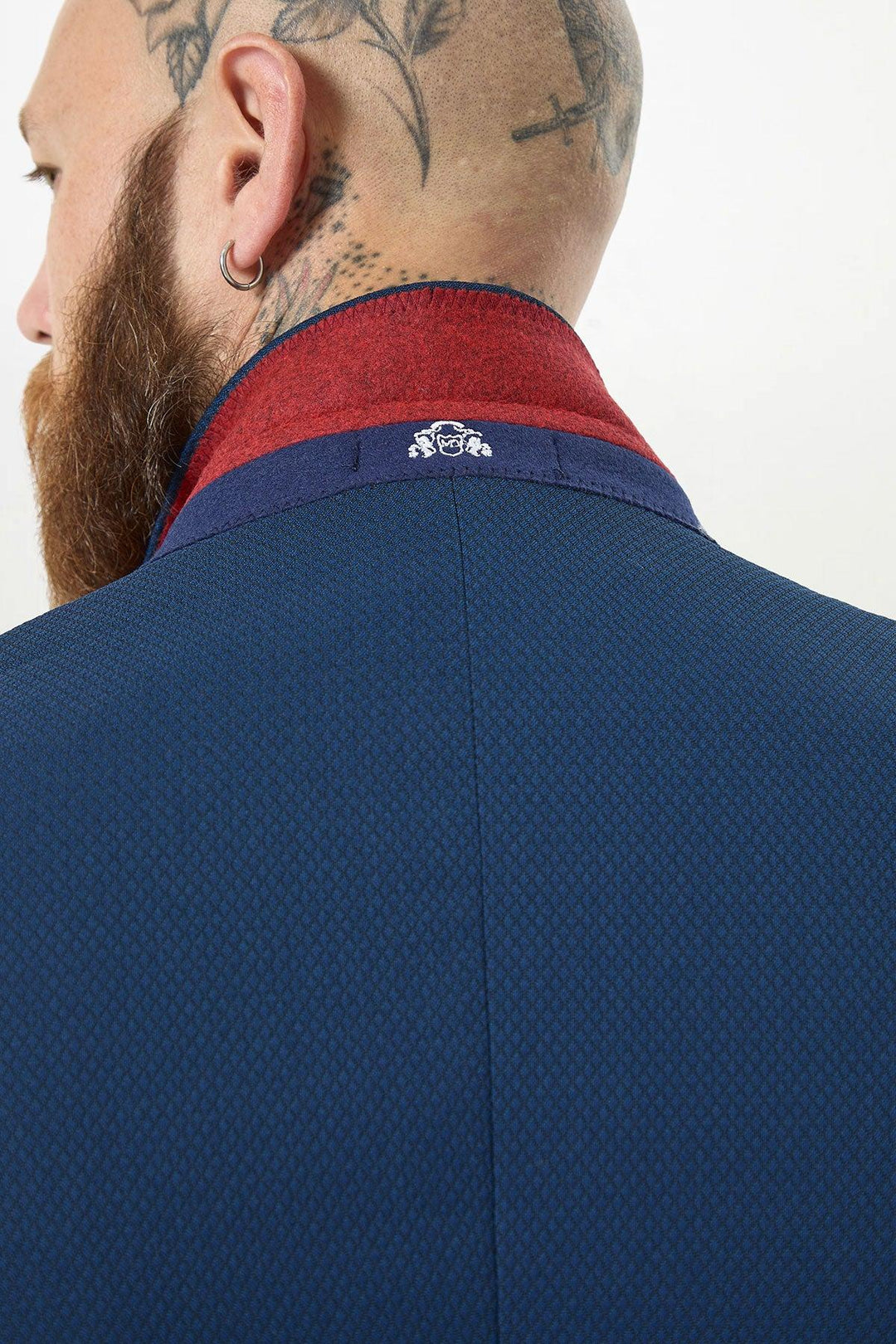 A man with a tattooed scalp, beard, and hoop earring is seen from behind, wearing the Marc Darcy MAX - Royal Blue Two Piece Suit with a red collar and an embroidered white lion crest under the collar.