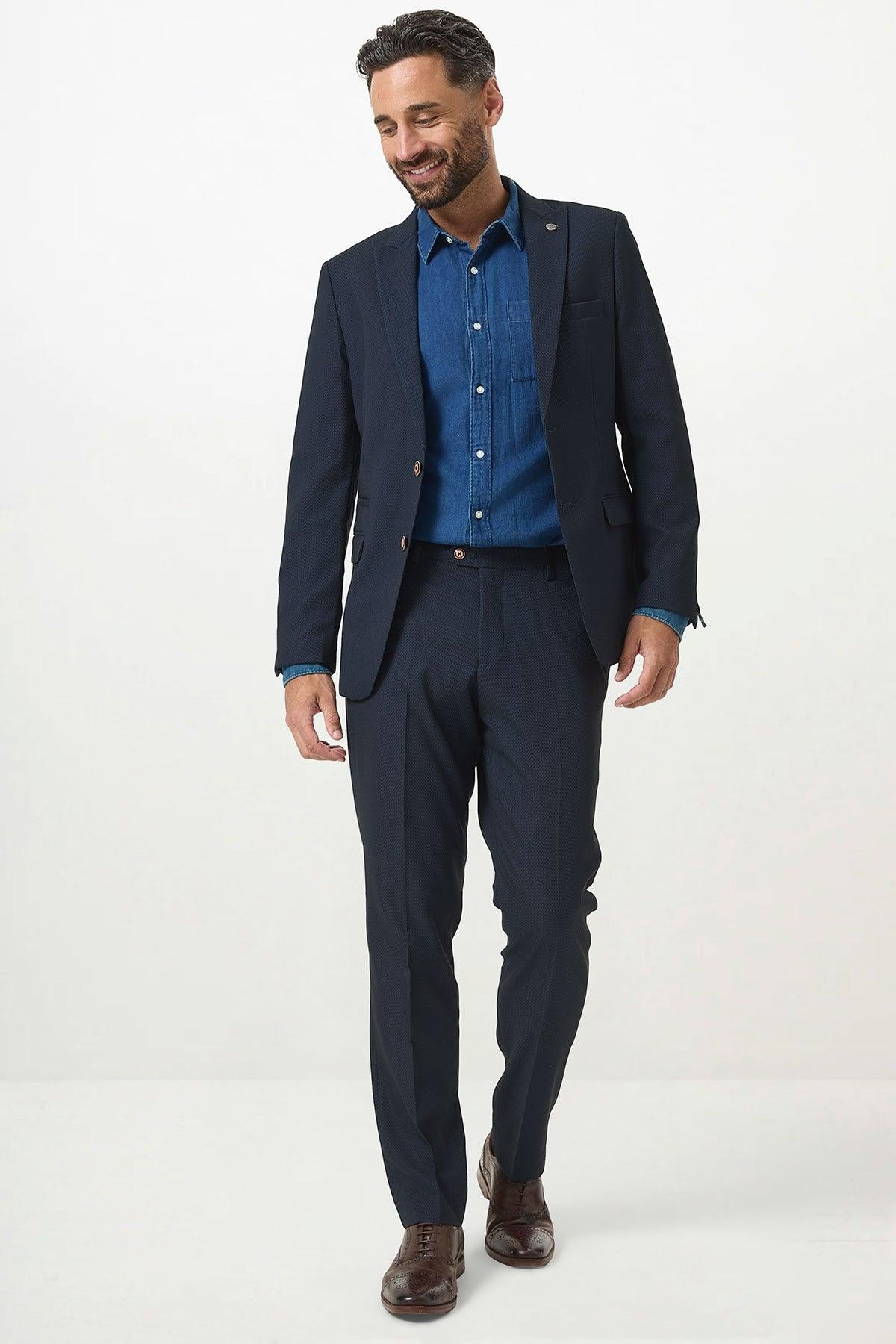 A man wearing the Marc Darcy MAX Navy Blue Two Piece Suit over a blue shirt with brown dress shoes stands smiling and looking down against a plain white background.