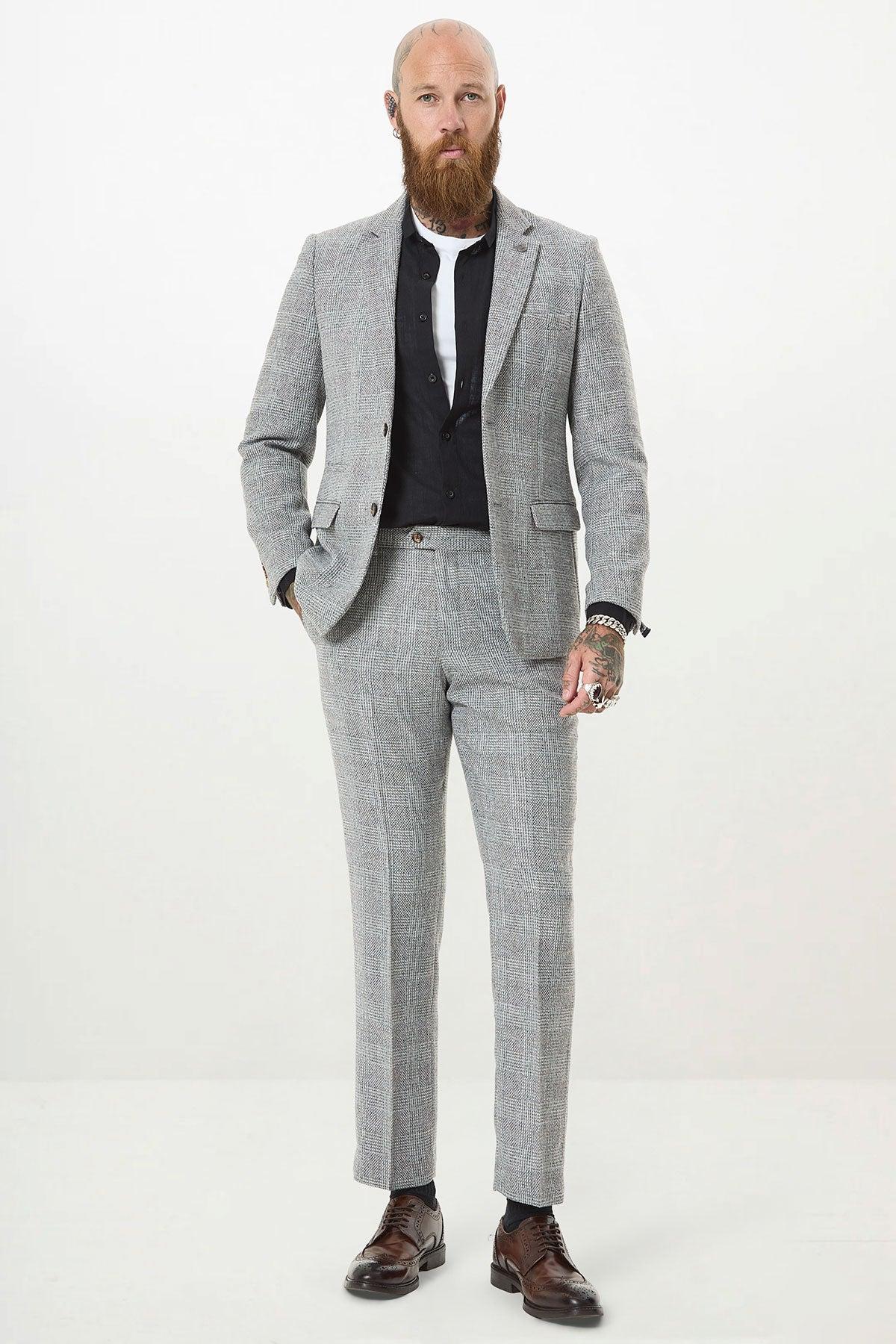 A bearded man with a tattooed hand stands against a white background in the marcdarcy KRIS Silver Grey Tweed Two Piece Suit, paired with a black shirt and brown dress shoes, looking serious with his hands at his sides.