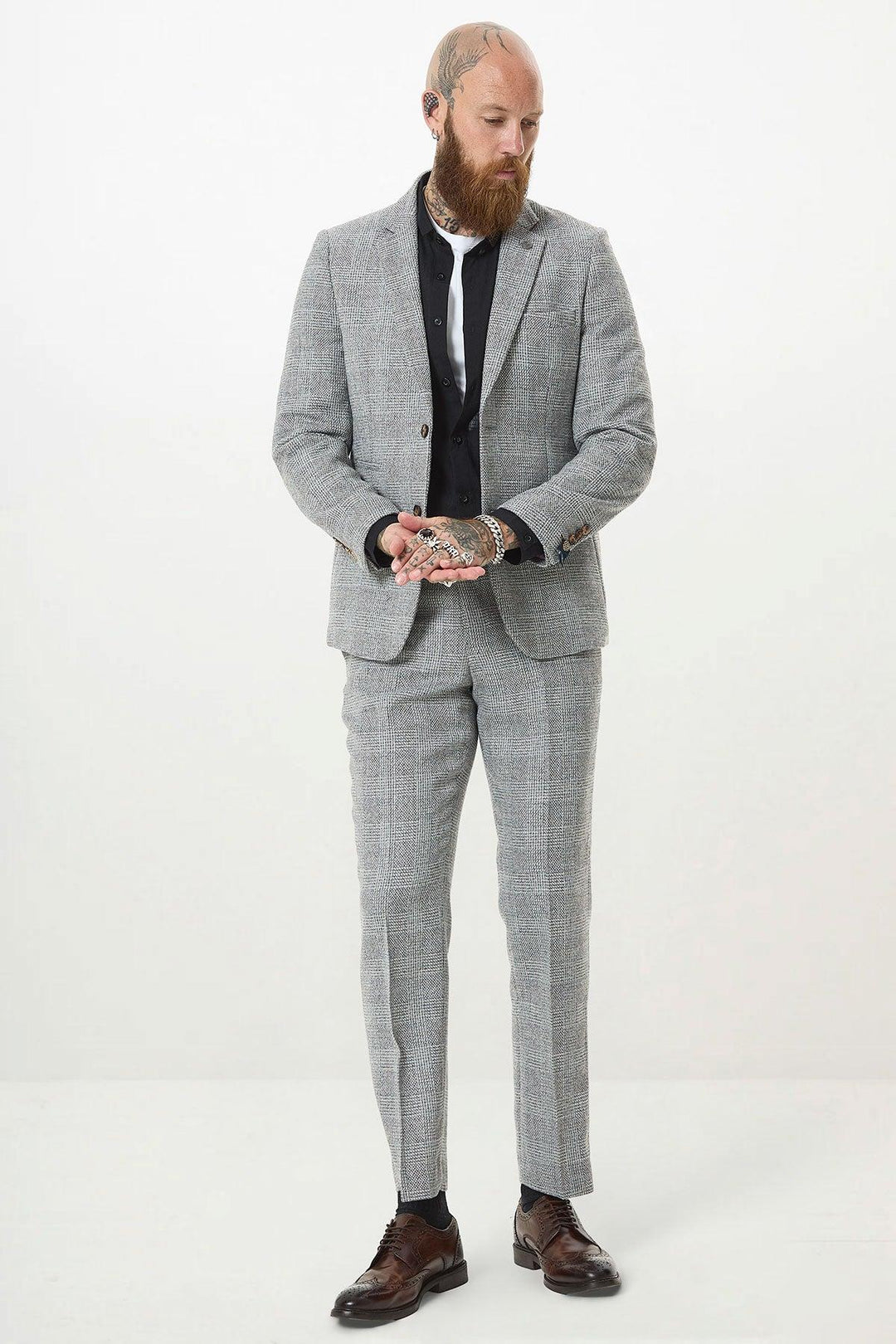 A bearded man with tattoos wears the marcdarcy KRIS - Silver Grey Tweed Two Piece Suit, paired with a black shirt and white tie, standing against a plain white background and looking down while clasping his hands.