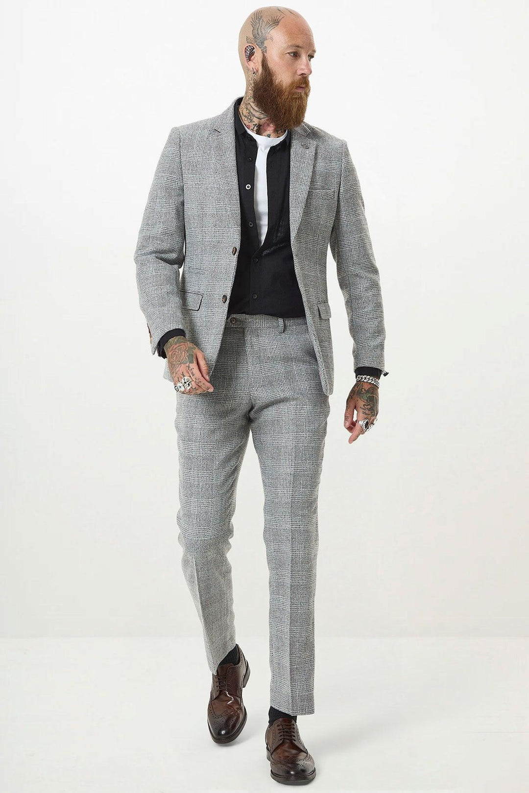 A bearded man with tattoos, dressed in the marcdarcy KRIS Silver Grey Tweed Two Piece Suit, white shirt, black waistcoat, and brown dress shoes, walks confidently against a plain white background.