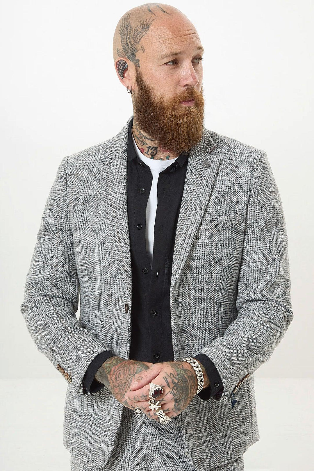 A bearded, tattooed man with a shaved head wears the marcdarcy KRIS Silver Grey Tweed Two Piece Suit over a black shirt, accessorized with rings and piercings. He stands with hands clasped, looking to the side against a plain background.