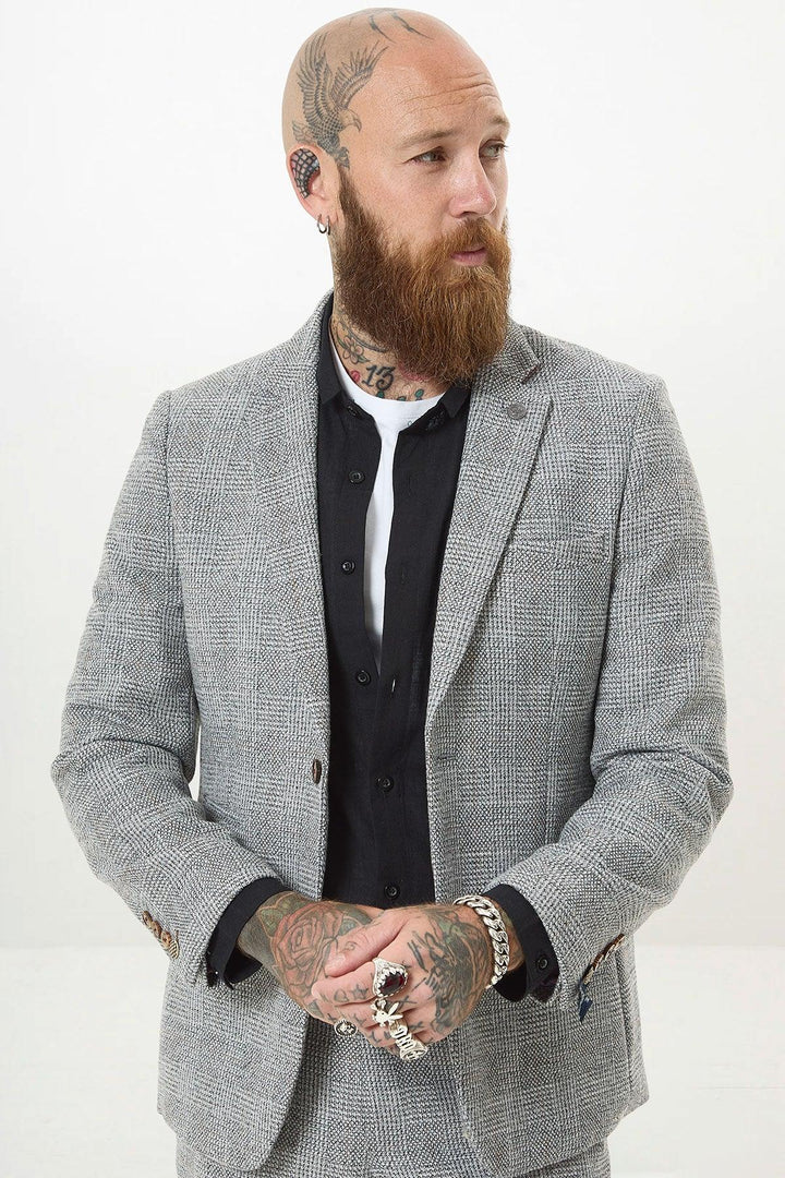 A bearded, tattooed man with a shaved head wears the marcdarcy KRIS Silver Grey Tweed Two Piece Suit over a black shirt, accessorized with rings and piercings. He stands with hands clasped, looking to the side against a plain background.