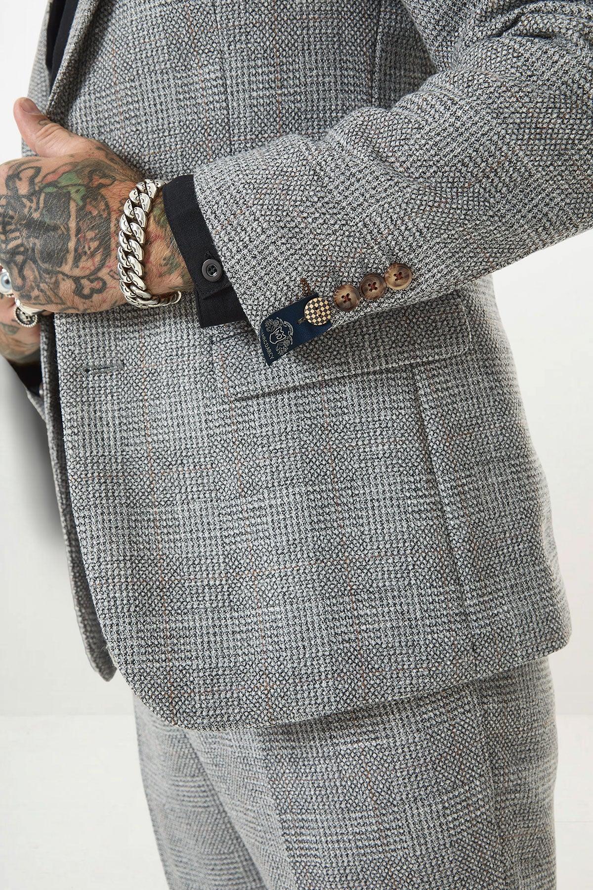 Close-up of a man in the marcdarcy KRIS Silver Grey Tweed Two Piece Suit with a Prince of Wales check, detailed buttons, and a branded tab. He pairs it with a black shirt and silver link bracelet.
