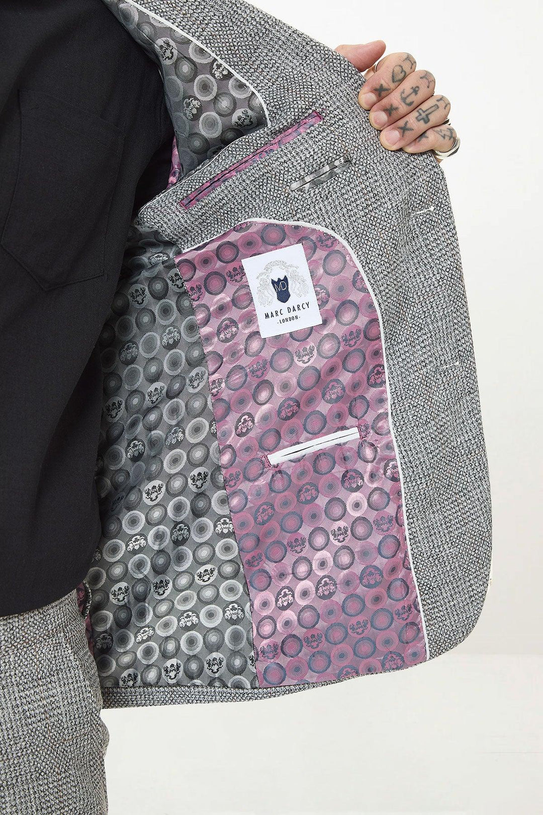 A person opens a KRIS - Silver Grey Tweed Two Piece Suit by marcdarcy, revealing a patterned lining with black and grey skulls on one side and pink on the other. The slim-fit blazer features a white NAKE ORACY label.