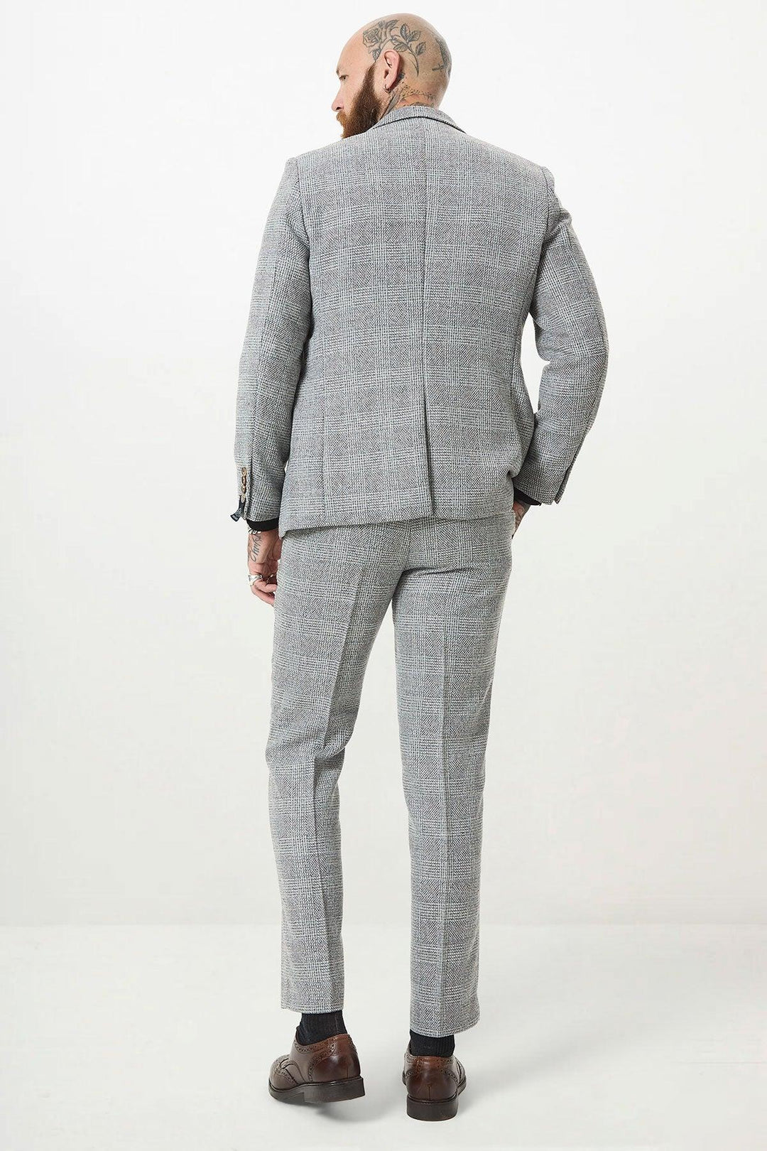 A man with head tattoos is facing away from the camera, wearing the marcdarcy KRIS Silver Grey Tweed Two Piece Suit with brown shoes and dark socks, against a plain white background.