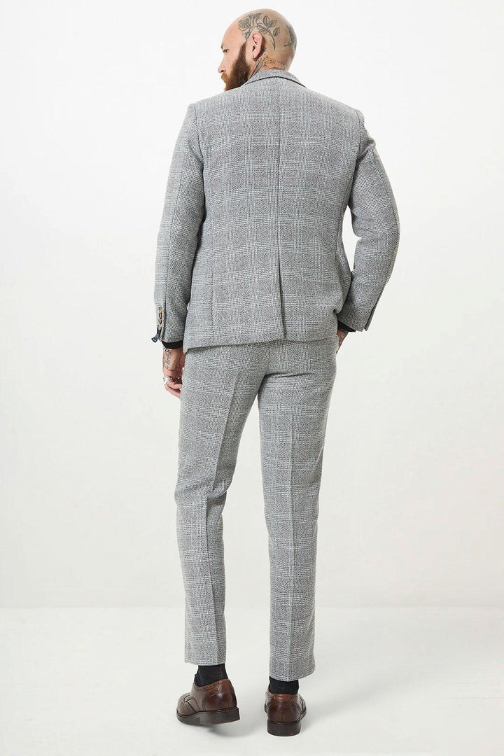 A man with head tattoos is facing away from the camera, wearing the marcdarcy KRIS Silver Grey Tweed Two Piece Suit with brown shoes and dark socks, against a plain white background.