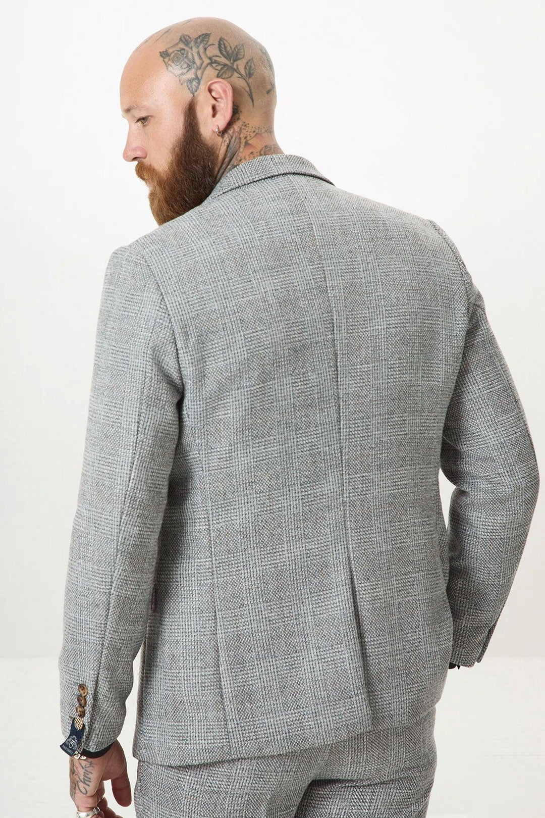 A man with a bald head, full beard, and head tattoos wears the marcdarcy KRIS Silver Grey Tweed Two Piece Suit. He faces away from the camera, glancing over his shoulder to highlight the suit’s back.