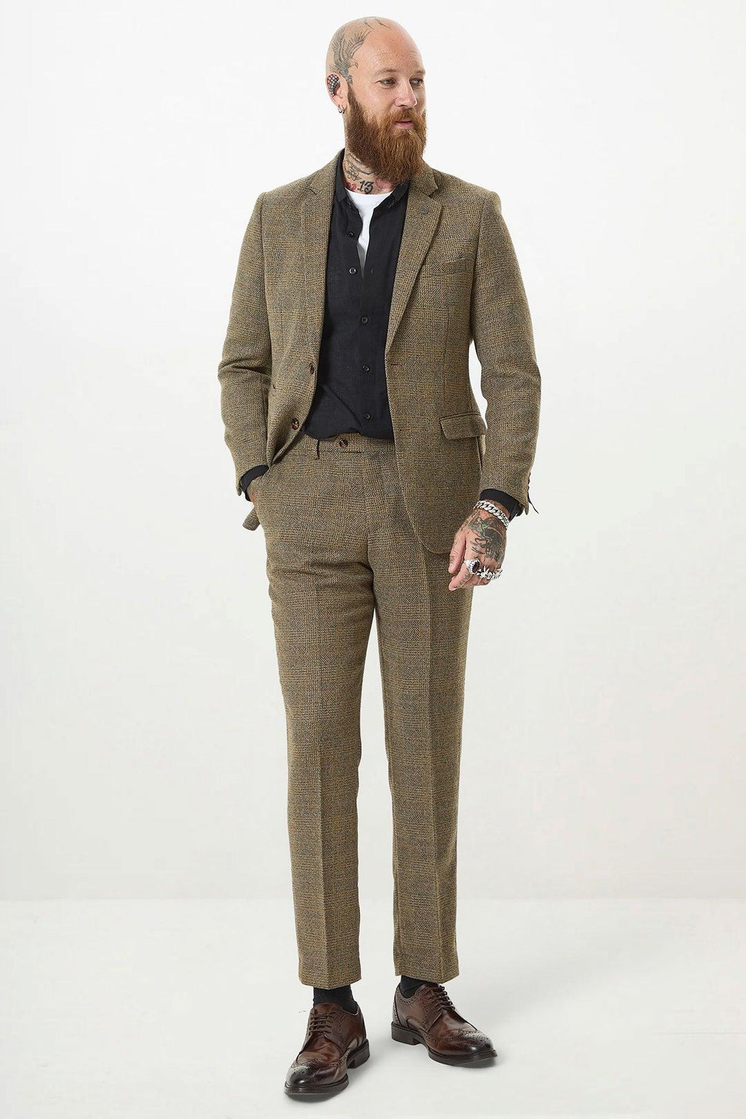 A bearded, tattooed man wears the marcdarcy KRIS Tan Tweed Two Piece Suit over a black vest and white shirt. He stands with hands in pockets against a plain white background, looking to the side and smiling slightly.