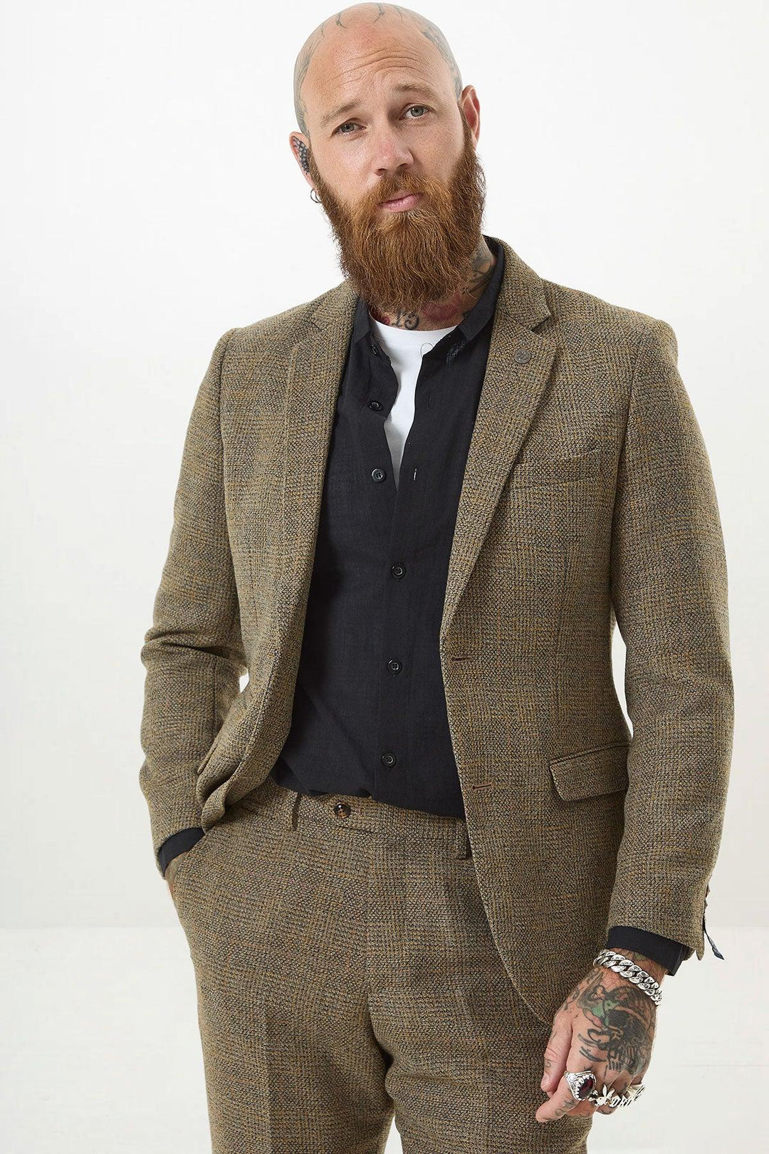 A bearded, tattooed man wears the marcdarcy KRIS Tan Tweed Two Piece Suit over a black shirt and white undershirt, standing against a plain white background with one hand in his pocket.