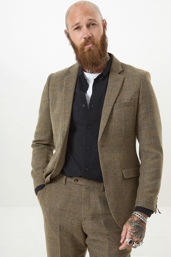 A bearded, tattooed man wears the marcdarcy KRIS Tan Tweed Two Piece Suit over a black shirt and white undershirt, standing against a plain white background with one hand in his pocket.