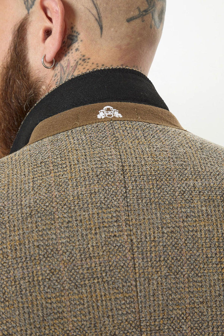 Close-up of the back of a person's neck wearing the marcdarcy KRIS Tan Tweed Two Piece Suit in Prince of Wales check with a black collar and white lion emblem. The person has a scalp tattoo, ear piercing, and short beard.