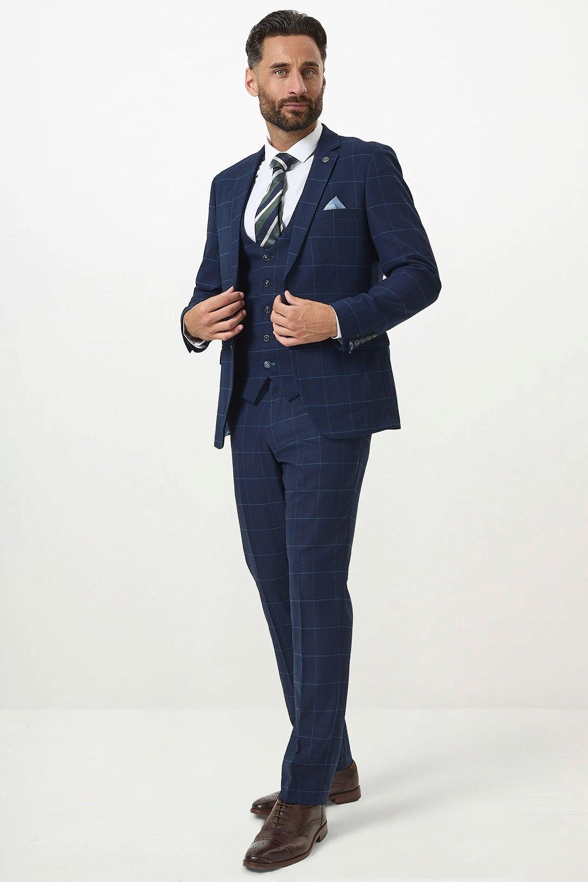 A man wearing the Marc Darcy EDINSON Navy Sky Check Three Piece Suit stands against a white background, adjusting his jacket over a white shirt, striped tie, and brown dress shoes while looking slightly to the side.